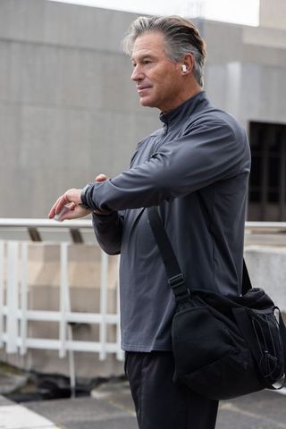 Mature man checking smartwatch on urban rooftop wearing earbuds and athletic jacket