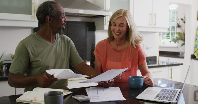 Senior couple sitting at kitchen table discussing finances with documents, laptop, coffee mugs around. Useful for themes related to retirement planning, budgeting, cooperation, financial management at home, elderly lifestyle, teamwork.