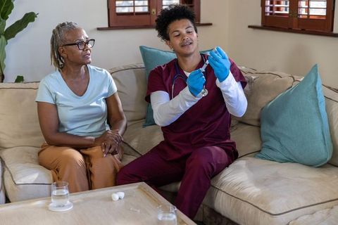Homecare nurse preparing vaccine syringe while senior african american woman watching on sofa