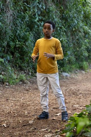 African american schoolboy standing on forest trail wearing mustard shirt and velcro sneakers