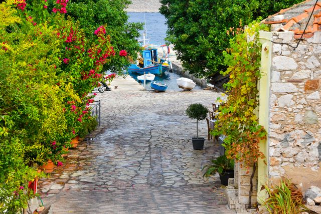 Narrow cobblestone alley with boats leading to a scenic beach in a coastal village. Vibrant greenery and colorful flowers add a touch of charm. Ideal for travel websites, summer vacation promotions, and brochures showcasing rustic Mediterranean destinations.