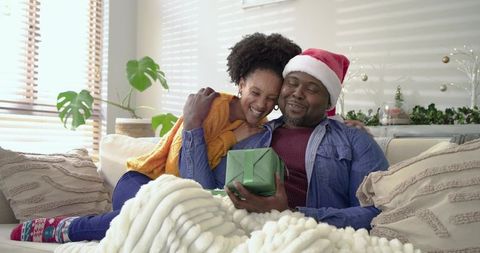 Cozy couple exchanging Christmas present on sofa under chunky blanket, holiday romance