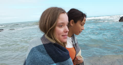 Friends Strolling by the Sea Sharing a Cozy Blanket