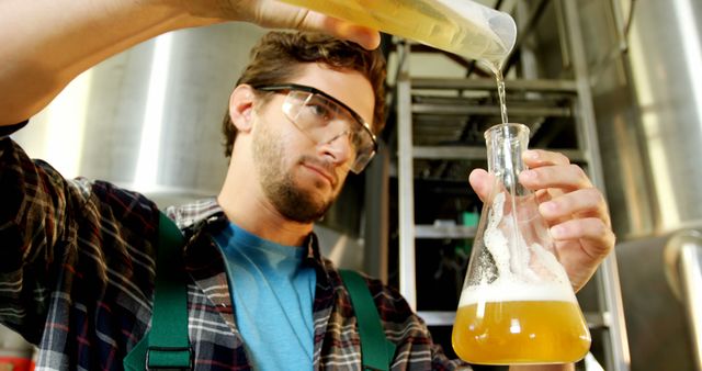 Microbrewer wearing protective goggles pouring craft beer into flask at brewery. Useful for content related to craft brewing, beer production, fermentation processes, and small-scale brewing facilities. Can be used in articles about the brewing industry, DIY beer making, and brewing science.