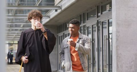 Urban graduate walking with friend holding coffee under glass canopy at campus entrance