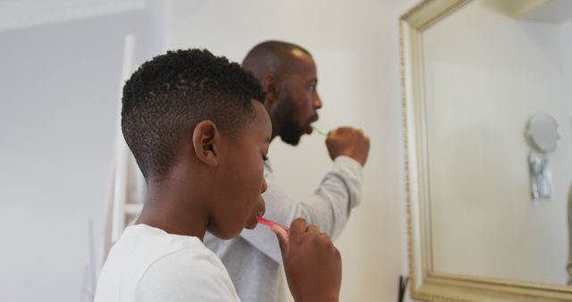 African american dad and son brushing teeth together looking in the mirror at home. fatherhood and love concept