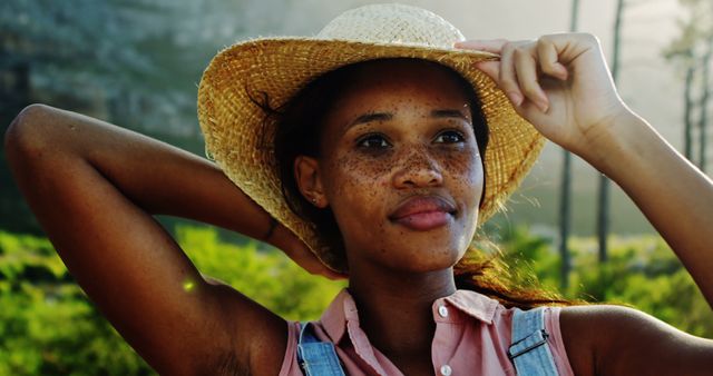 Young African American woman enjoys a sunny day outdoors, with copy space. She exudes confidence and relaxation, tipping her straw hat in a serene environment.