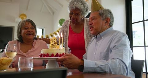 Elderly friends are celebrating a birthday in a cozy living space. Two women and one man are joyfully gathered around a cake adorned with glowing candles. They are wearing festive attire and a party hat is in view, emphasizing the celebratory theme. There are decorations like balloons enhancing the festive atmosphere. This image can be used for promoting senior activities, lifestyle articles focusing on friendships among elderly individuals, and party planning for older adults.