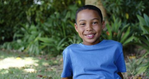 Smiling african american boy sitting on grass in sunlit garden wearing blue t-shirt