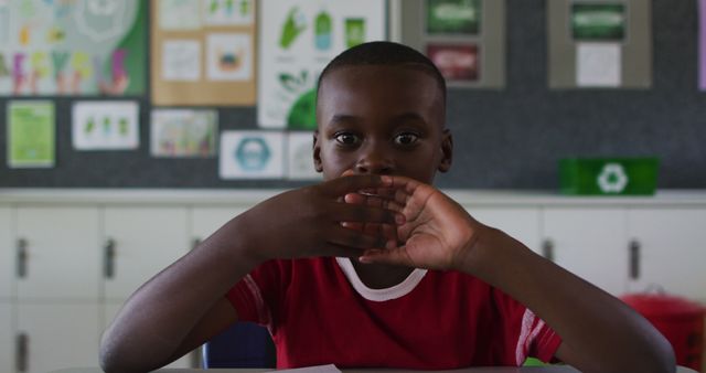 Young boy sitting in classroom, seems to be thinking while covering mouth with hands. Ideal for use in educational contexts, psychological studies, or back-to-school promotions. Showcases child engagement and thoughtful moments in school setting.