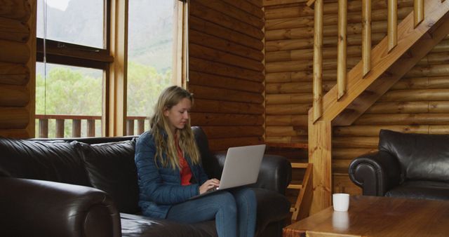 Young woman sitting on a sofa with a laptop in a cozy wooden cabin. Ideal for themes related to remote work, nature retreats, modern technology in natural settings, and ergonomic home offices.