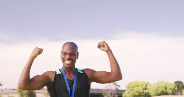 Young male athlete smiling and raising arms in celebration of victory outdoors. Ideal for illustrating sports success, motivation, endurance, and healthy lifestyles.