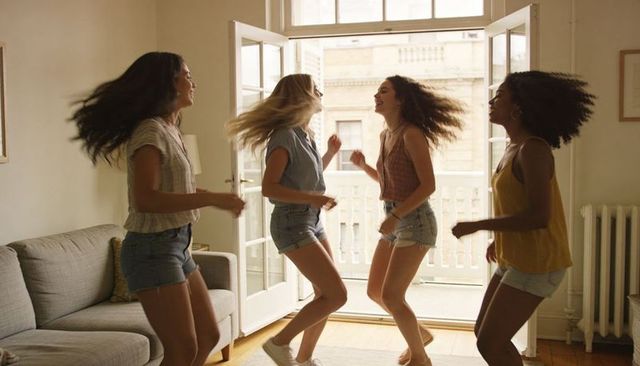 Four friends dancing and jumping in sunlit apartment near open french doors
