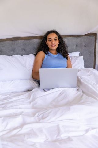 Woman working on laptop in bed leaning against tufted headboard remote work lifestyle
