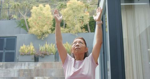Senior asian woman stretching arms upward on patio wearing smartwatch outdoor wellness