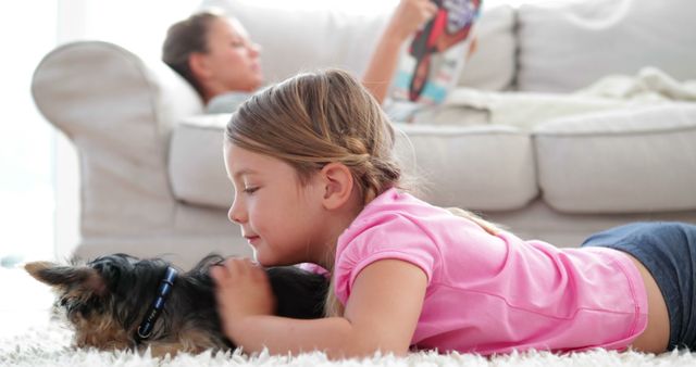 Young girl lies on a soft carpet, happily petting a small dog. Background shows a comfortable sofa with another person resting, adding to the warm, family atmosphere. Useful for themes related to family life, pet companionship, and home relaxation.