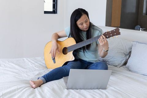 Asian woman practicing acoustic guitar on bed following laptop for online music lesson