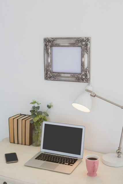 This image depicts a modern workspace featuring a laptop, books, a coffee mug, a smartphone, a desk lamp, and a decorative frame on a white wall. Ideal for use in articles or advertisements related to home office setups, productivity tips, modern decor, and technology in daily life.