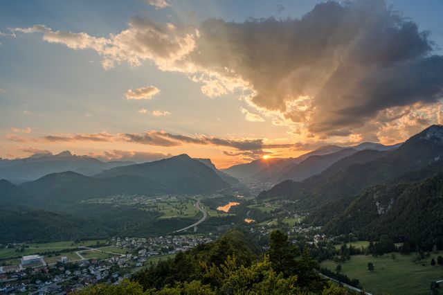 Sunset over mountains with rays piercing through clouds, casting a golden hue on the lush valley below. Ideal for travel advertisements, nature and landscape photography, desktop wallpapers, and promotional material for adventure tourism.