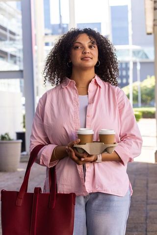 Young african american professional carrying coffee cups and red tote in sunlit urban plaza