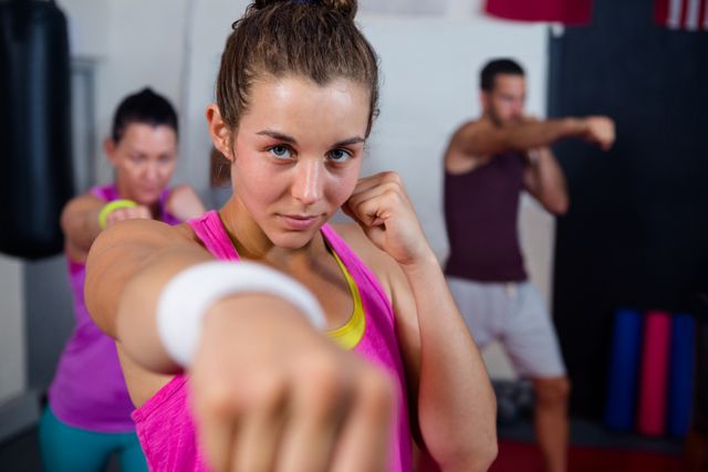 Young woman practicing boxing in a fitness studio with two other individuals in the background. Ideal for use in articles or advertisements related to fitness, boxing training, women's empowerment, and healthy lifestyles.
