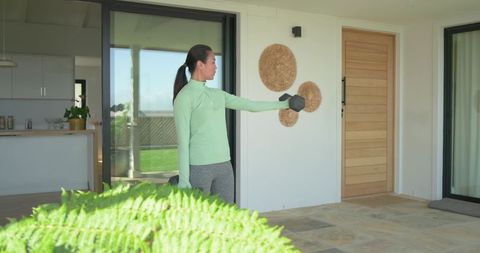 Asian woman lifting hex dumbbell on sunny patio in mint top and grey leggings