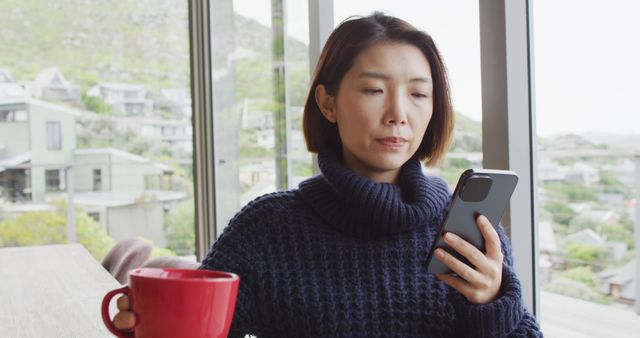 Woman sitting indoors holding a red coffee mug and using a smartphone. She appears focused on the mobile screen, suggesting she is reading or messaging. Light-filled room with a cozy atmosphere. Suitable for concepts related to daily life, technology use, morning routines, and casual home activities.