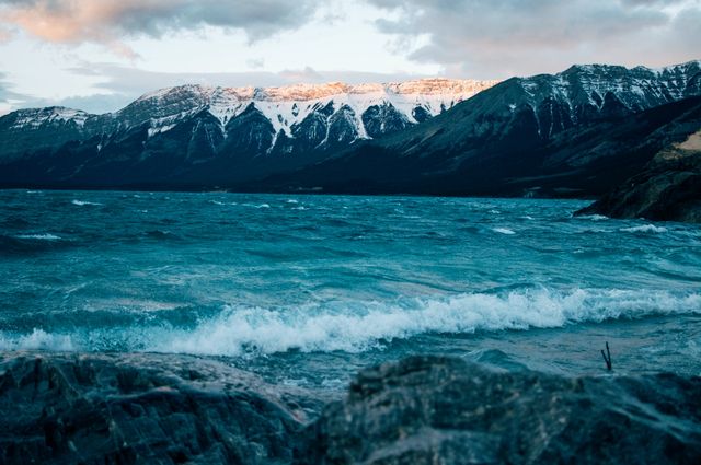 Turquoise waves crash against rocky shore with snow-capped peaks in background under overcast sky. Perfect for travel blogs, adventure promotions, nature documentaries, and landscape posters.