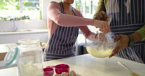 Multicultural Couple Whisking Batter Together in Sunlit Home Kitchen Wearing Striped Aprons