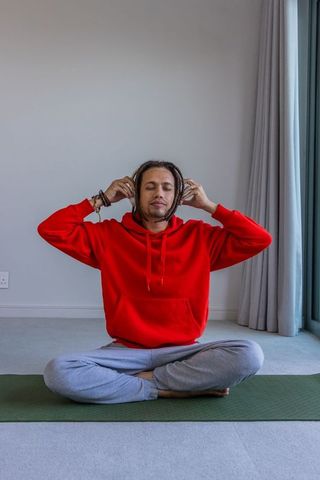 African american man meditating with headphones on green yoga mat in bright living room