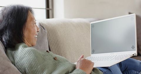 Senior Asian woman relaxing on beige couch using white laptop, smiling and typing at home