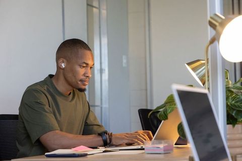 African american man working on laptop at modern desk with earbuds, lamp and plants