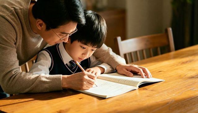 Father guiding son through homework at wooden table, focused tutoring and family learning