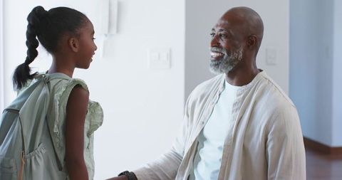 African american father kneeling and greeting daughter with mint backpack in modern home entryway