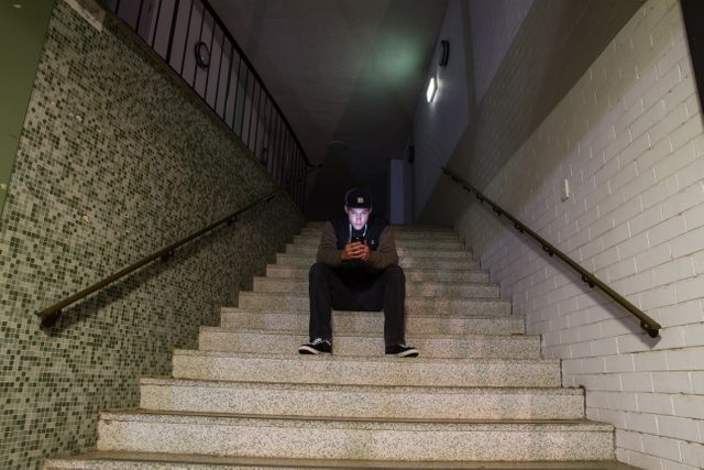 Young man sits alone on stairway in dimly lit corridor, absorbed in his phone. Ideal for themes related to technology, youth, solitude, night-time, or social media usage. Suitable for use in articles, blog posts, ads, or social media campaigns focusing on modern lifestyle, isolation, or tech consumption habits.
