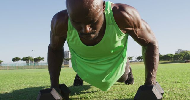 An athletic man in a neon green tank top doing push-ups with dumbbells outdoors on a grass field. This image can be used for fitness blogs, exercise tutorials, sports advertisements, or health and wellness promotional materials.