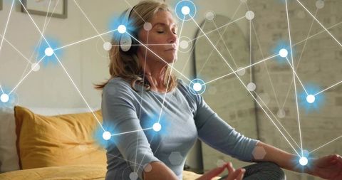 Senior woman meditating with headphones on bed with glowing neural network overlay
