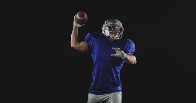 American football player wearing blue jersey and helmet, captured mid-throw on black background. Perfect for sports promotions, athletic advertising, and fitness campaigns.