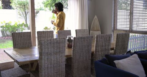 Man Standing by Sliding Door Drinking Coffee While Holding Phone in Sunlit Dining Room