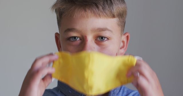 Boy holding yellow face mask in front of his face. Concept of health protection, safety, and measures against viral diseases. Perfect for educational content on health safety, pandemic-related articles, and child care topics.