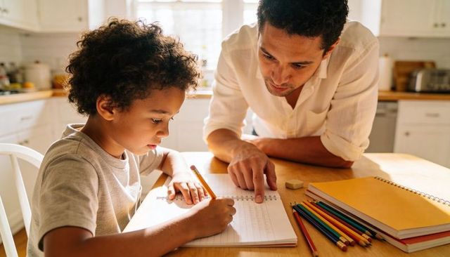 Father guiding child doing homework at kitchen table with notebooks, pencils, warm light
