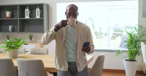Mature african american man sipping coffee, checking phone in bright modern dining room