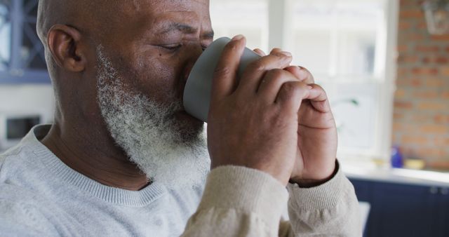 Elderly man with a grey beard drinks coffee in a cozy kitchen with natural light. Ideal for content related to morning routines, retirement life, staying at home, and relaxation. Useful for articles, advertisements, or blogs focused on senior well-being, lifestyle, and health.