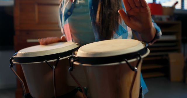 Person playing bongo drums indoors with close-up on hands hitting drums, showcasing rhythm and percussion. Suitable for music-related content, educational materials on percussion instruments, or promotional materials for music events.