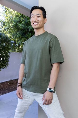 Asian man leaning on column wearing olive t-shirt and smartwatch smiling on sunlit patio