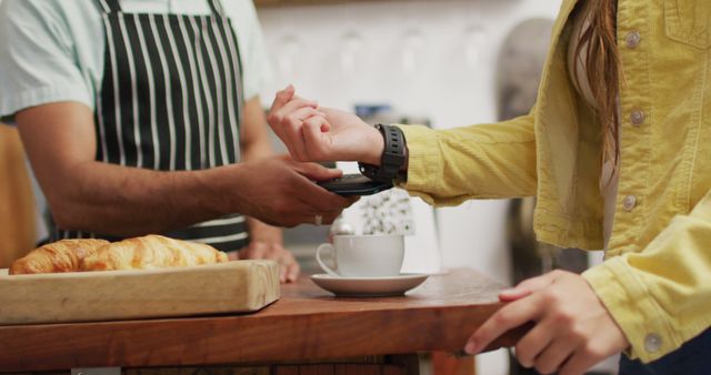 A customer wearing a yellow jacket uses a smartwatch to make a cashless payment at a cafe. The male cashier in an apron scans their watch. There is a coffee cup and a croissant on the table. This visual is ideal for illustrating modern payment methods, convenience in retail, advancements in technology, and lifestyle articles focusing on digital transactions.