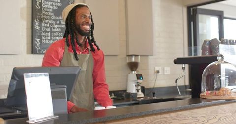 Smiling african american barista serving coffee behind counter tapping pos touchscreen