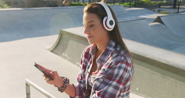 Smiling caucasian woman wearing headphones, using smartphone and waving at a skatepark. hanging out at skatepark in summer.