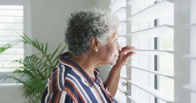 Senior woman stands by window, gazing outside with pensive expression. Background includes bright natural light and potted plant. Ideal for themes of aging, contemplation, loneliness, and reflection.