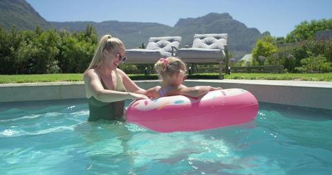 Mother teaching daughter on pink donut float wearing sunglasses in sunny backyard pool
